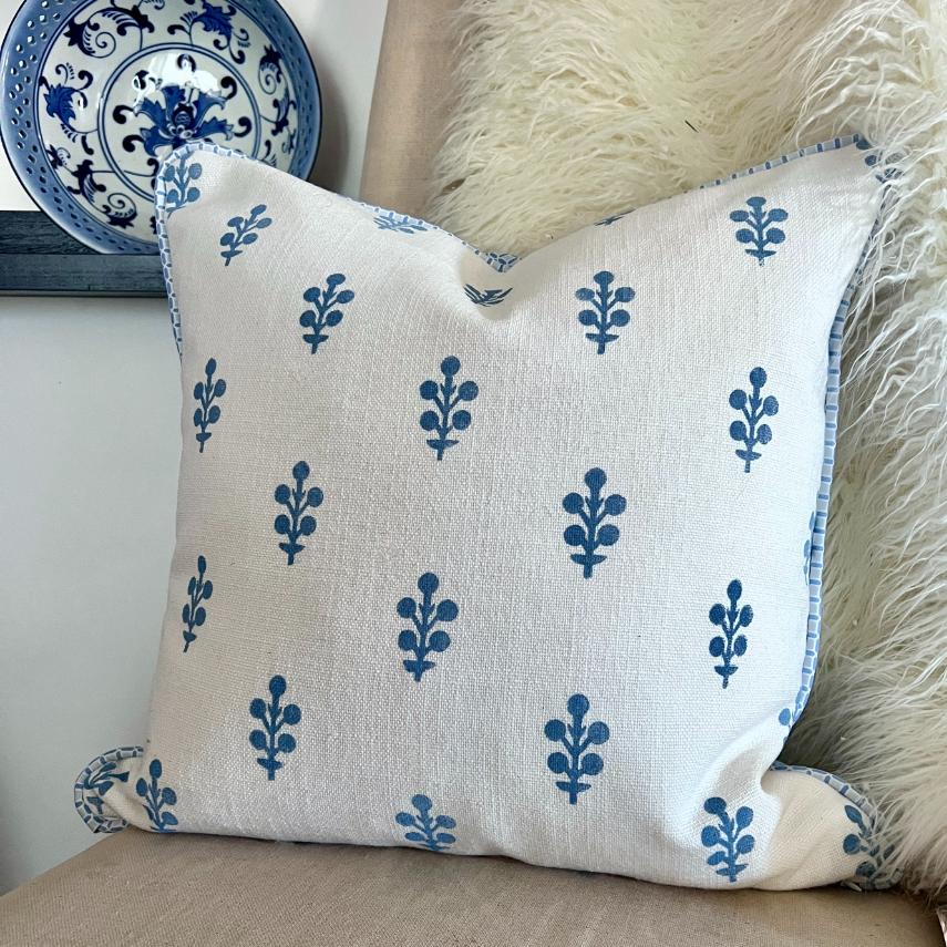 A white cushion with blue leaf motifs sits on a beige chair. The cushion has a blue and white striped edge, and a furry cream throw is draped behind it. A decorative blue and white plate is visible in the background.