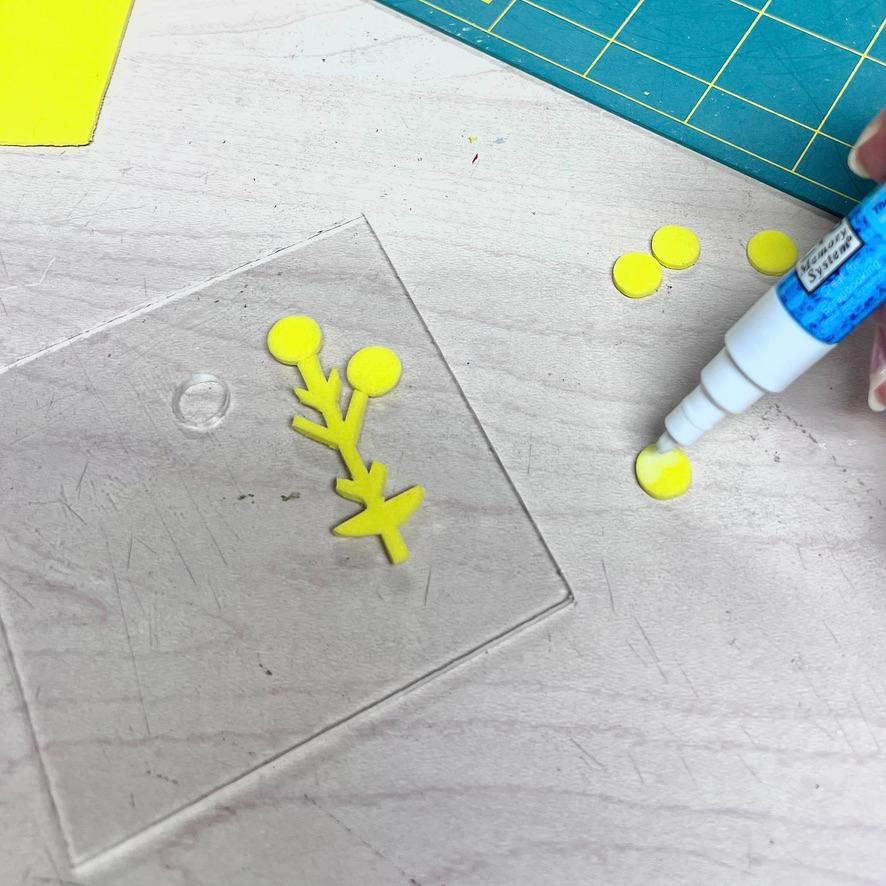A hand uses a blue pen to color a small yellow circle next to a clear square with a yellow paper plant shape on a white table. A green cutting mat and a yellow sheet are in the background.