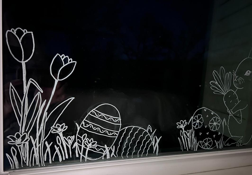 White line drawings on a window show flowers, decorated Easter eggs, and a bunny holding a large carrot, all set in grass. The scene is seen at night with a dark background outside.