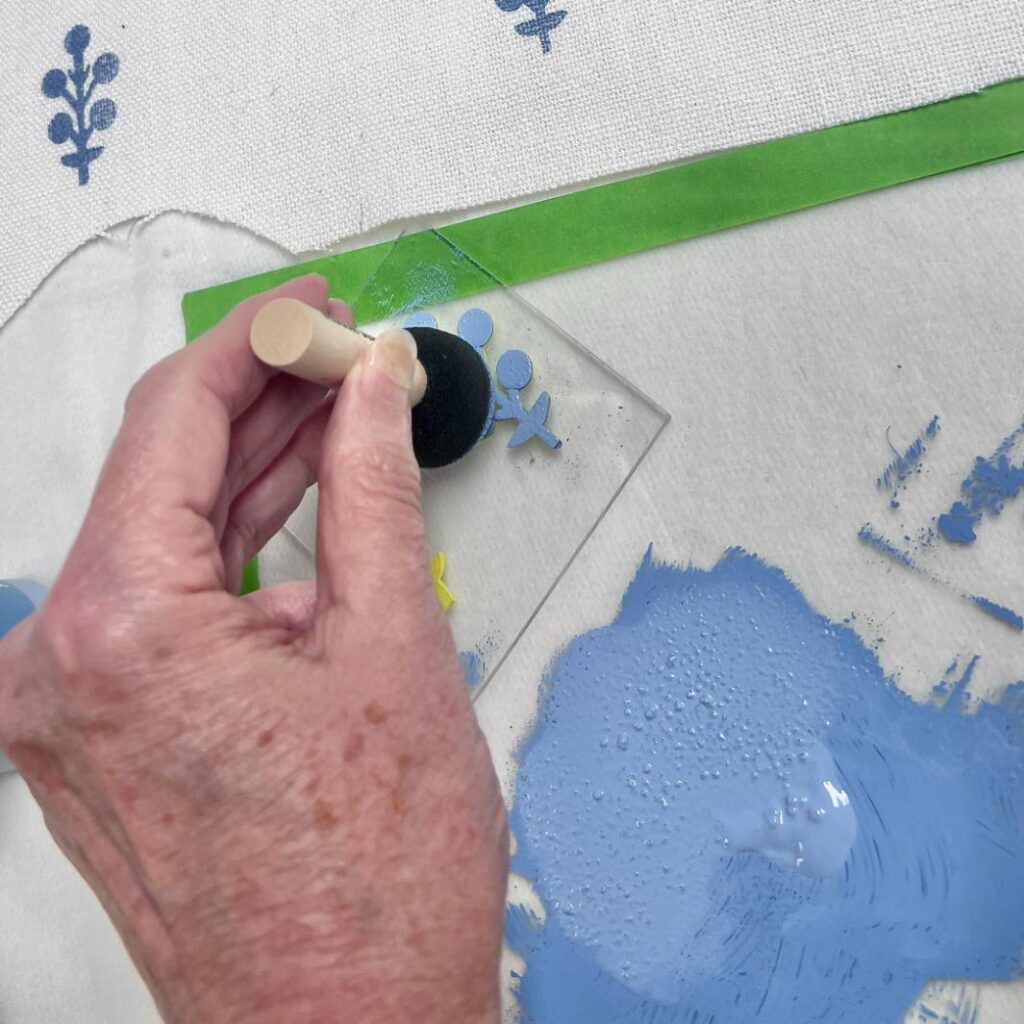 A hand uses a sponge dabber to apply blue paint to a stencil shaped like a person, placed on a white fabric with green painter’s tape and blue painted designs nearby.