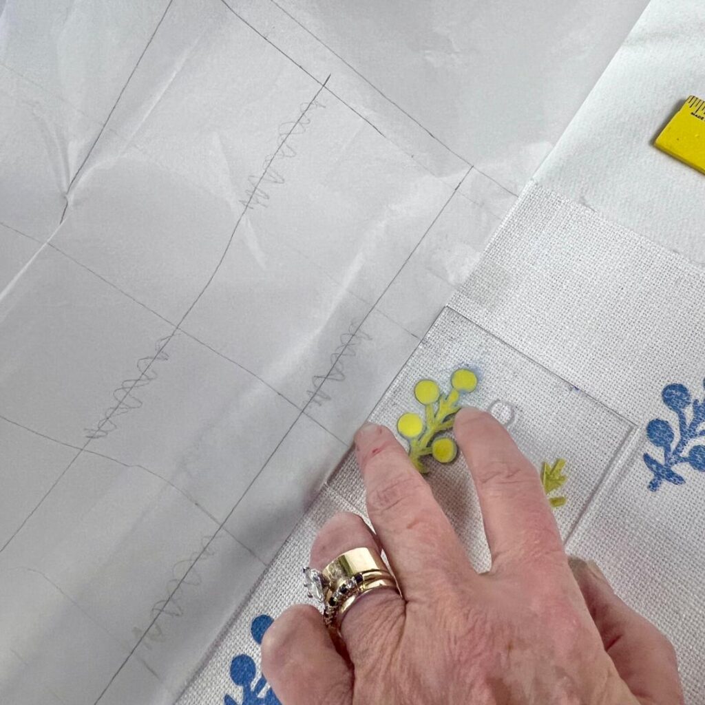 A hand with rings holds a clear acrylic template with a yellow and green floral design over fabric with blue printed flowers, next to sewing pattern paper with zigzag lines. A yellow ruler is partially visible in the corner.