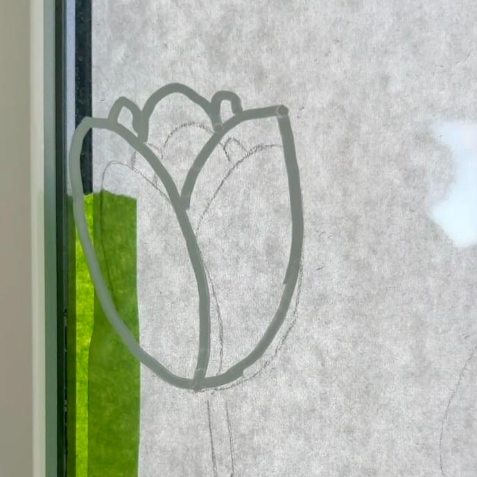 A simple outline of a tulip flower is drawn on a window with a green vertical strip taped beside it. Light filters through the window in the background.