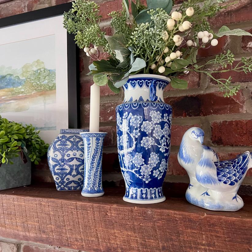 A wooden mantel displays blue and white ceramics, including a vase with white and green flowers, two patterned vases, and a decorative bird, set against a brick wall and framed artwork.