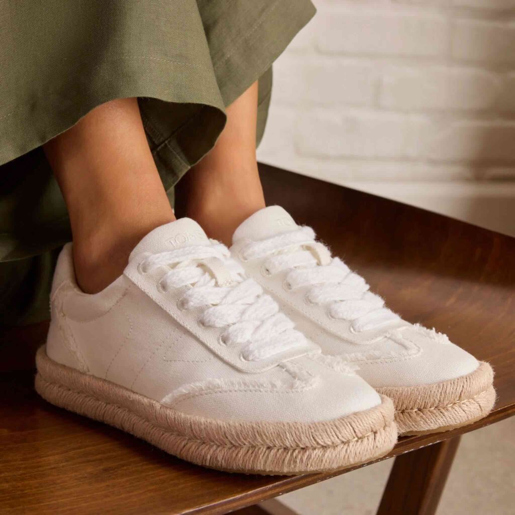 A person wearing white canvas sneakers with frayed edges and thick, woven jute soles, sitting on a wooden surface. The person is dressed in olive green pants.