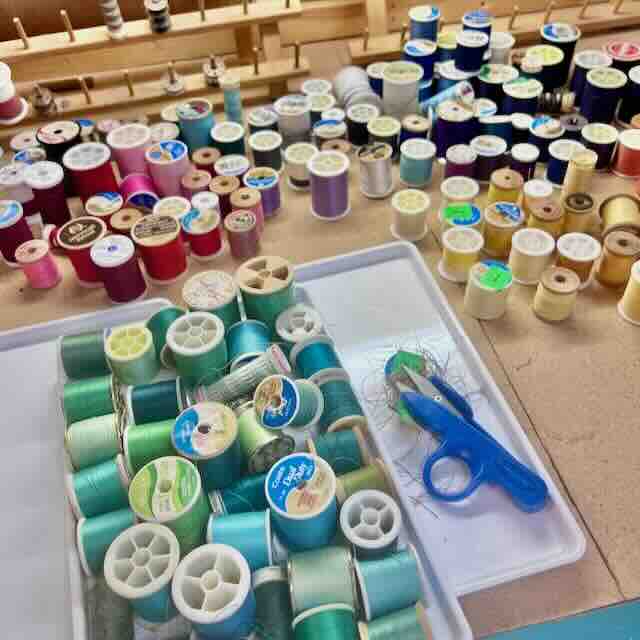 Spools of thread in various colors are arranged on a table, with a focus on greens and blues in the foreground. A blue pair of scissors lies beside the threads on a white tray.