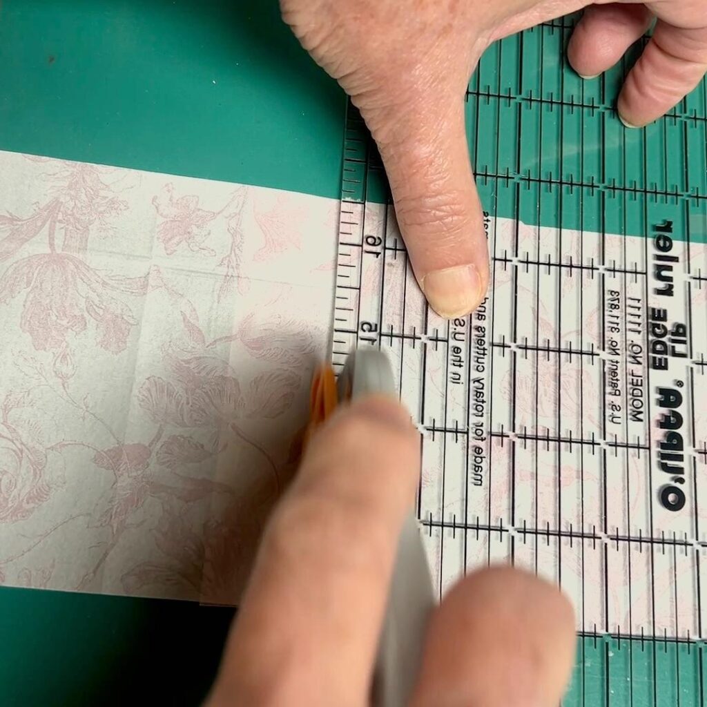 A person uses a rotary cutter and a clear ruler to cut patterned paper on a green cutting mat, carefully aligning the paper for a precise cut during their thrifted basket makeover project.