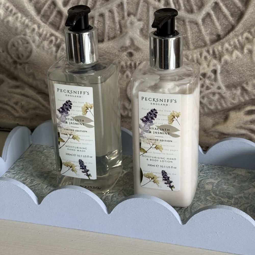 Two bottles of Pecksniff’s England Hyacinth & Jasmine hand wash and hand lotion with pump dispensers sit on a light blue, scalloped functional basket tray, against a textured, patterned background.