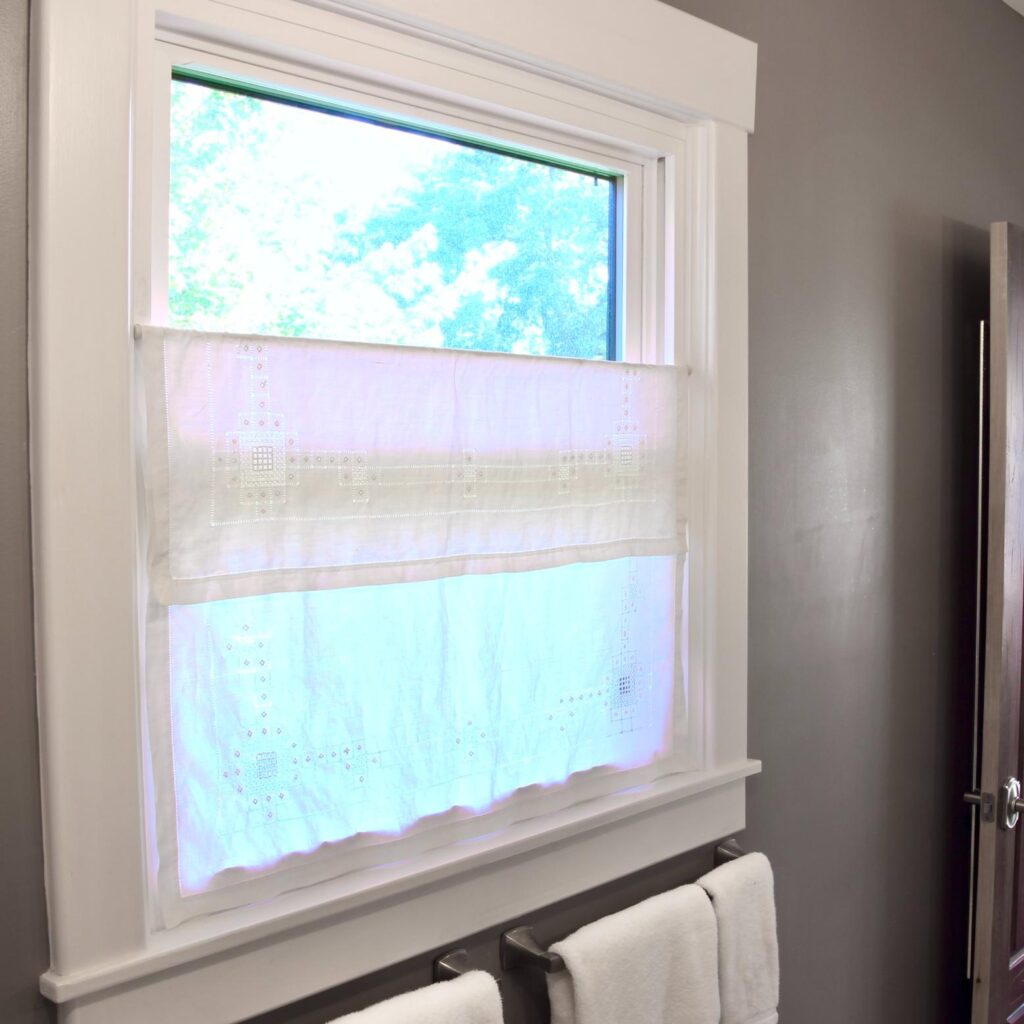 A window with white embroidered café curtains hangs on extra long curtain rods in a bathroom with gray walls. Sunlight pours through the window, and two white towels are neatly placed on a rack below.