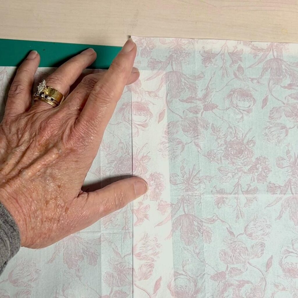 A hand with rings is folding floral patterned paper near a green edge on a light-colored surface, crafting details for a thrifted basket makeover.