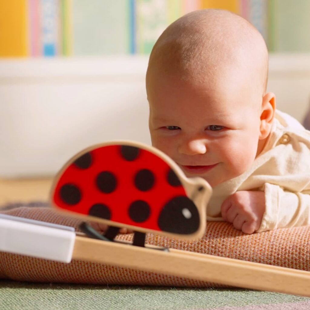 A baby lying on their stomach smiles while looking at a wooden toy ladybug with red and black spots on a wooden base indoors.