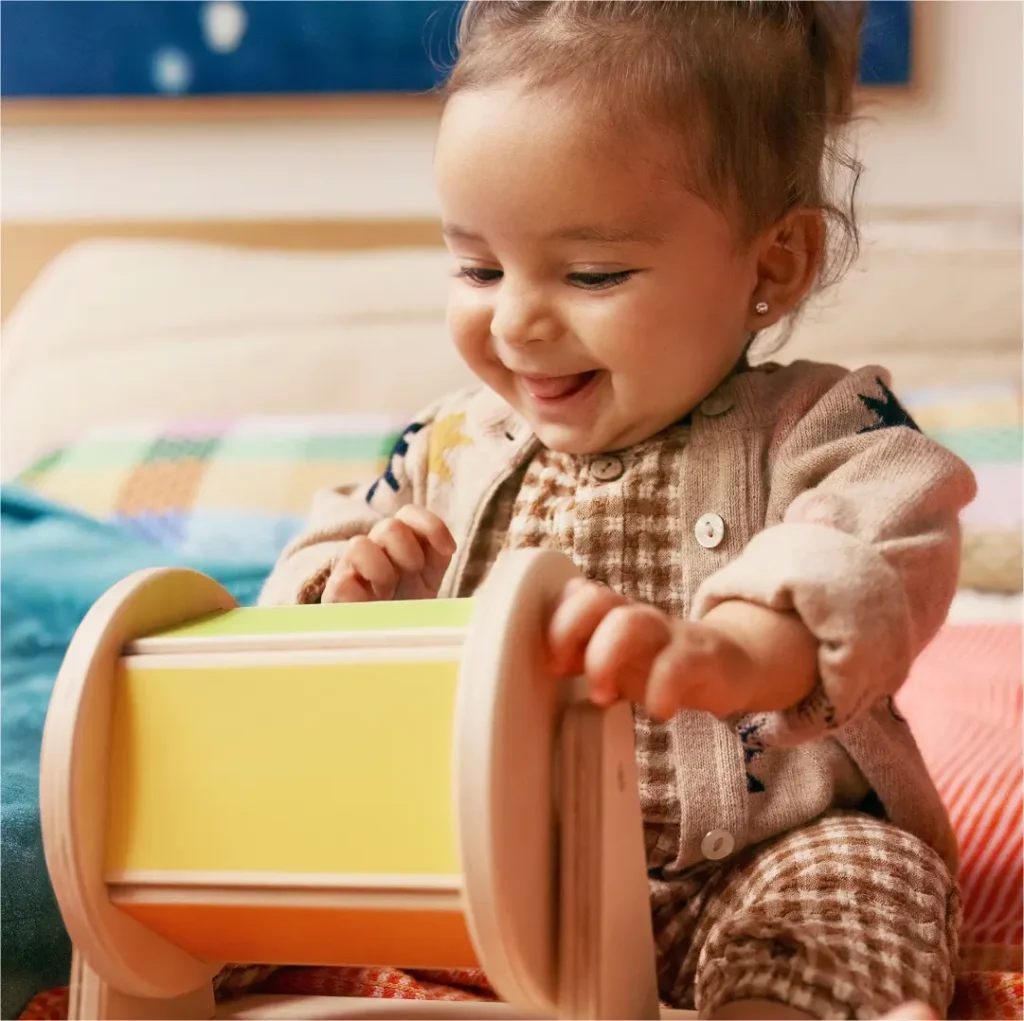 A smiling baby in a cozy outfit plays with a colorful wooden drum toy on a bed, surrounded by soft, patterned blankets.