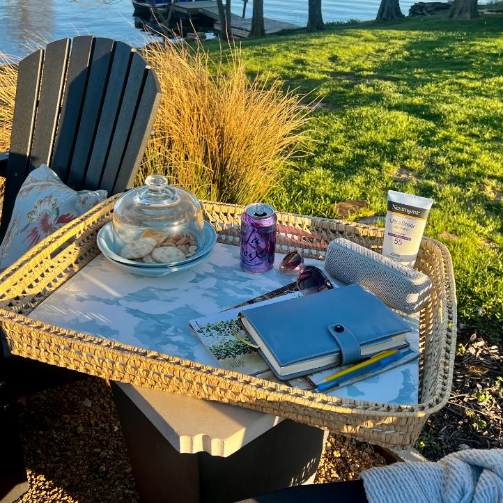 A functional basket tray on an outdoor table holds cookies under a glass dome, a can of sparkling water, sunglasses, sunscreen, a notebook with pens, and a rolled towel—all set against a serene lakeside view.