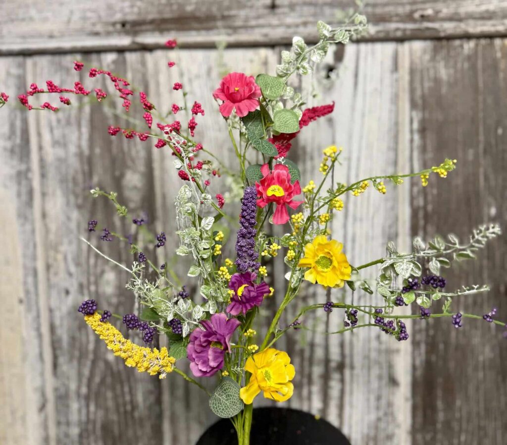 A colorful bouquet of artificial flowers, including red, purple, and yellow blooms with green leaves, is arranged in a black vase in front of a weathered wooden background.