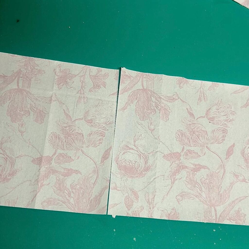 Two pieces of light-colored paper with a pink floral pattern are placed side by side on a green surface, with a small gap between them—perfect for crafting projects like a thrifted basket makeover.