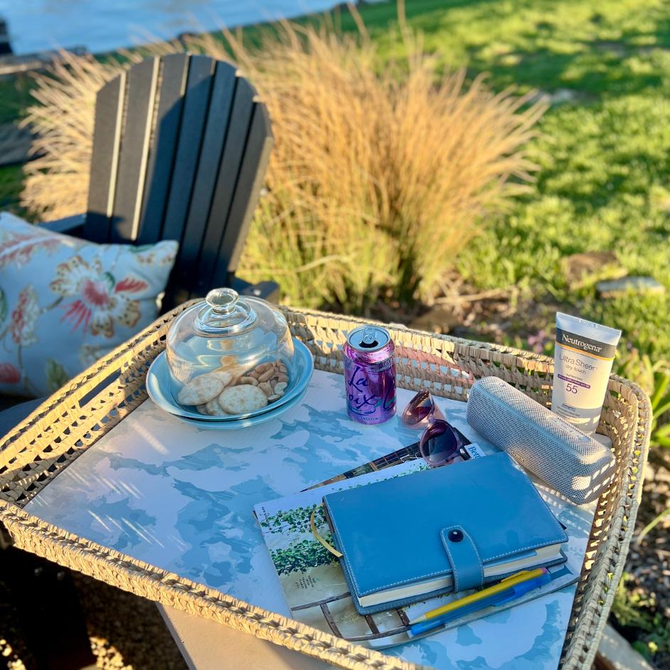 A Functional Basket Tray on an outdoor table holds cookies under a glass dome, a planner, magazines, pens, sunglasses, a rolled towel, LaCroix, and sunscreen. A black chair and grass with tall plants complete the inviting scene.