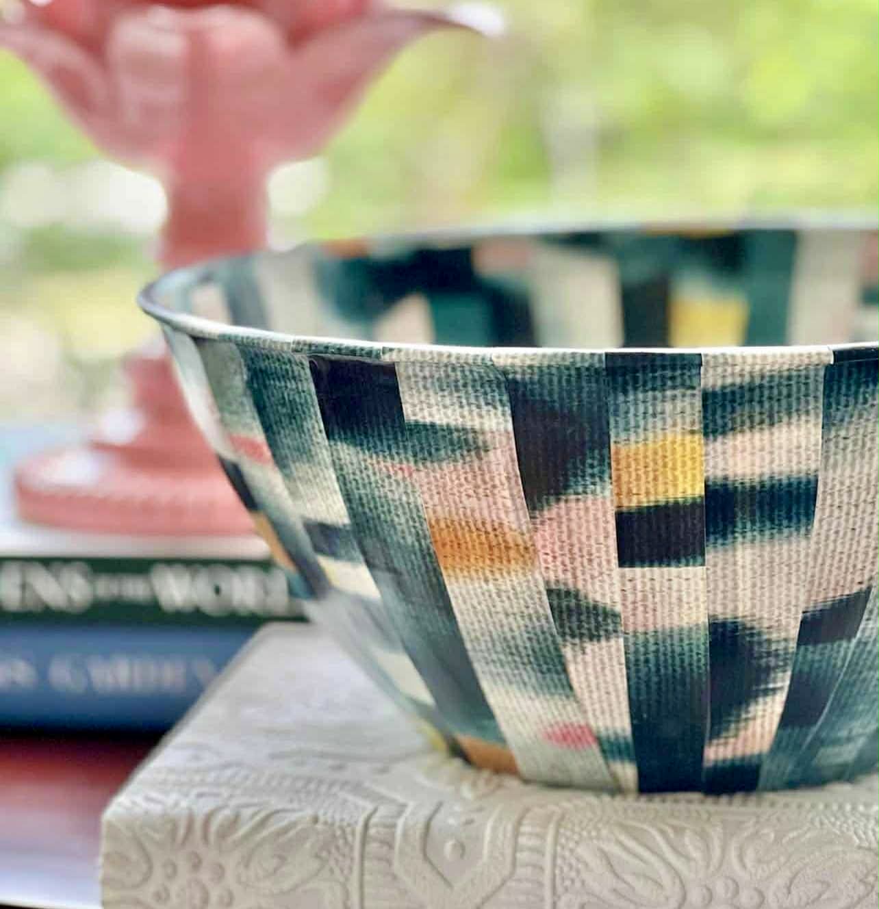 A colorful, patterned bowl sits on a textured white cloth atop a functional basket tray, with a stack of books and a pink decorative object blurred in the background. The scene is softly lit with a natural, green backdrop.