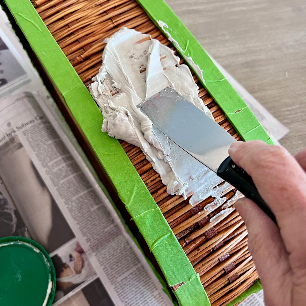 A hand uses a putty knife to spread white spackle or plaster over a section of a thrifted basket, bordered by green painter’s tape, with newspaper underneath for protection during this creative makeover.