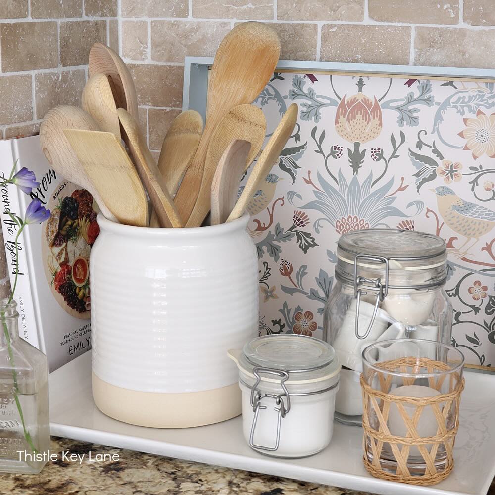 A kitchen countertop displays a white ceramic jar holding wooden utensils, two glass jars with white ingredients, a small glass with wicker cover, a cookbook, and a flower in a bottle, all arranged on a functional basket tray with a tile backsplash.