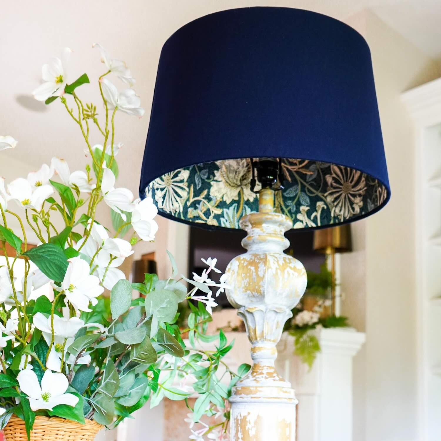 A rustic, distressed wooden lamp with a dark blue lampshade featuring a floral pattern on the inside, next to a bouquet of white flowers and green leaves arranged in a functional basket tray.