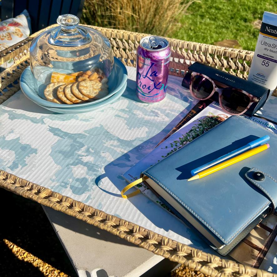 A functional basket tray holds crackers under a glass dome, a can of LaCroix, sunglasses, Neutrogena sunscreen, a blue notebook with a yellow pen, and magazines—all set outdoors in sunlight on the table.
