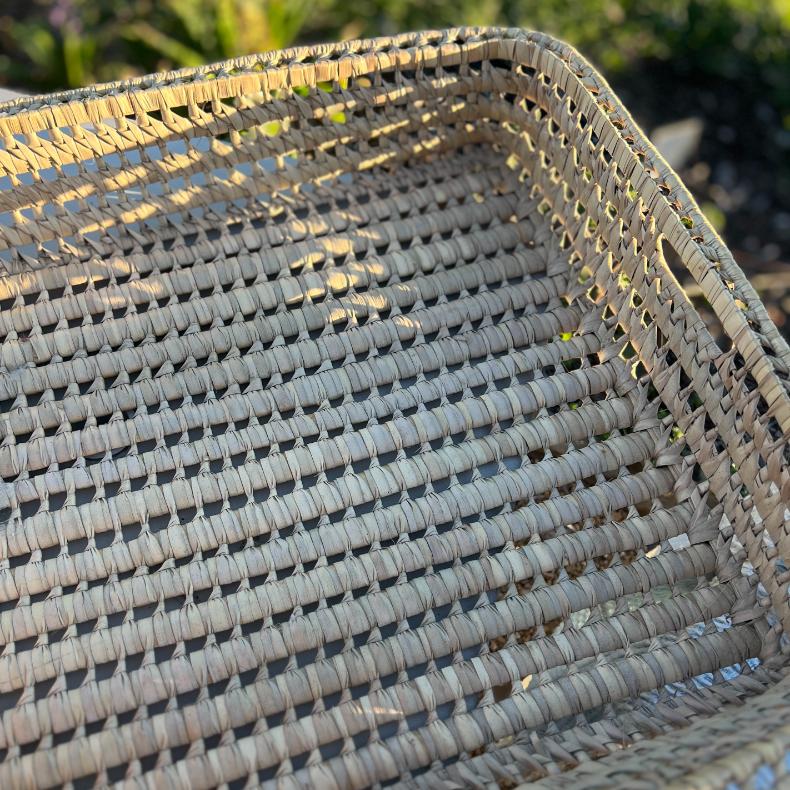 A close-up of a rectangular, light brown wicker basket with a woven pattern, seen outdoors in sunlight. This functional basket tray has handles on the sides and features visible shadows from its textured weave.