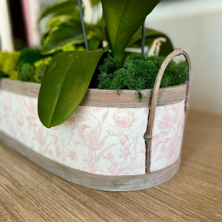 A close-up of a thrifted basket makeover, transformed into a decorative planter with pink floral patterns, filled with green moss and leafy plants, sitting on a wooden surface.