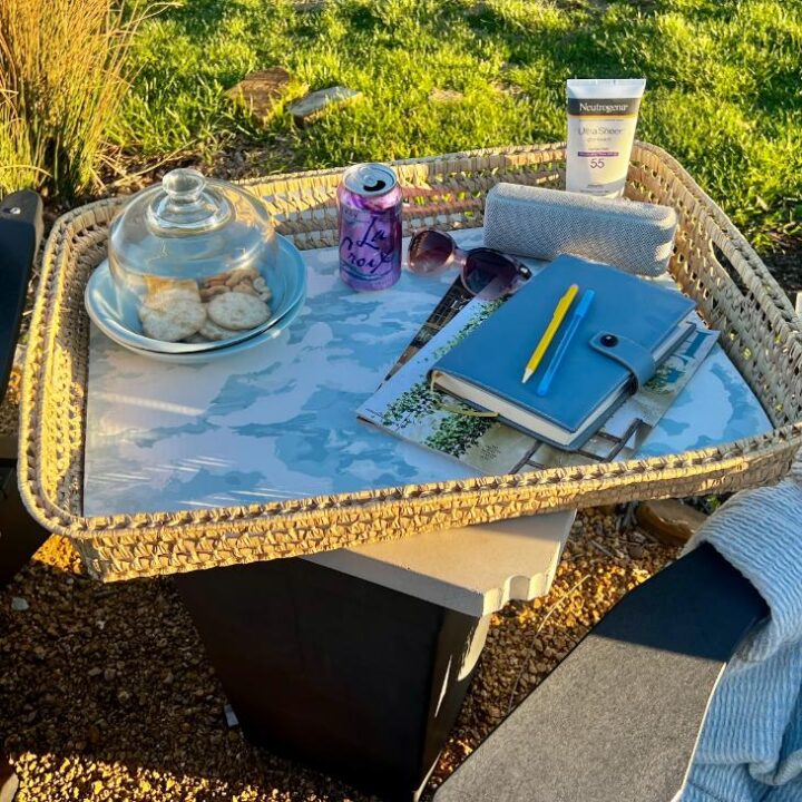 A functional basket tray on a small outdoor table holds a plate of crackers under a glass dome, a can of sparkling water, sunglasses, a rolled towel, sunscreen, notebook, pen, and blue planner. Grass is visible in the background.