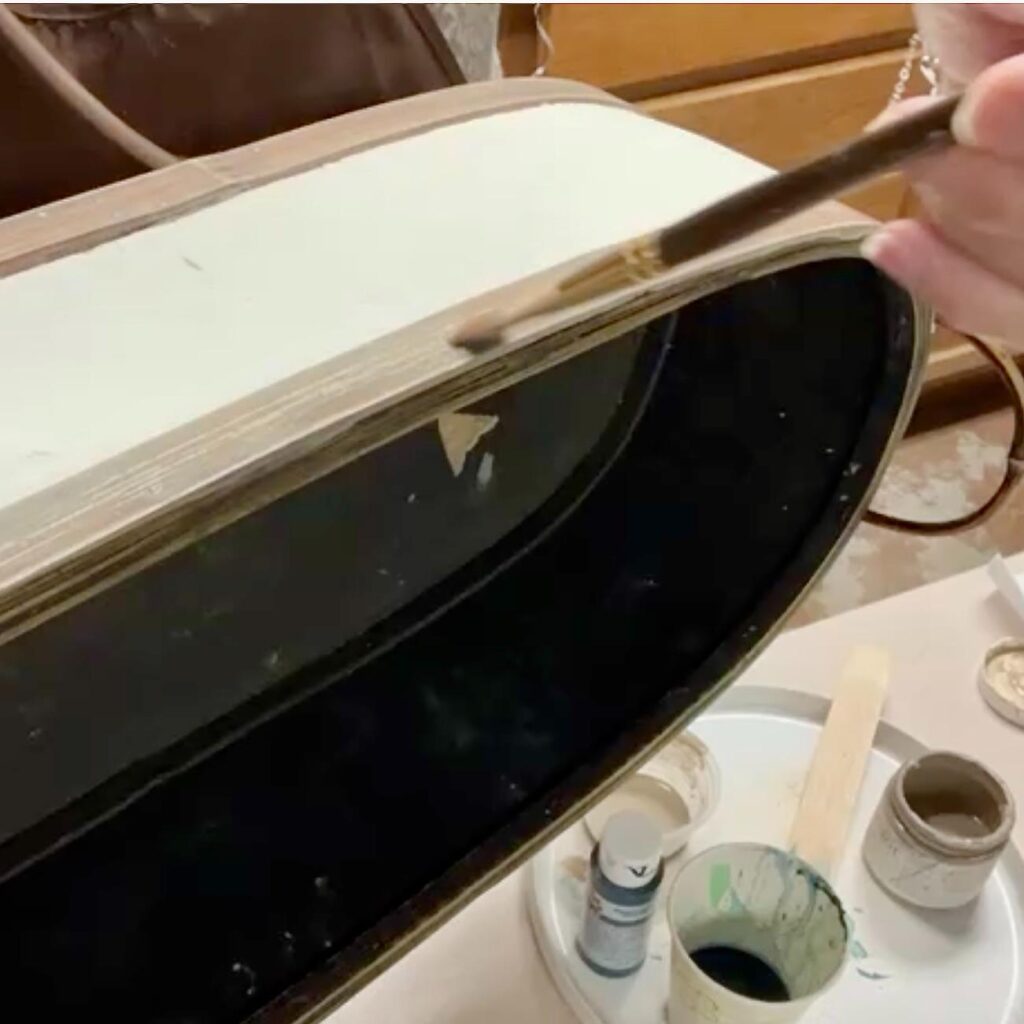 A hand holds a paintbrush, applying paint to the edge of a curved wooden object in a thrifted basket makeover. Nearby, there are paint supplies including jars, a palette, and a wooden stick.