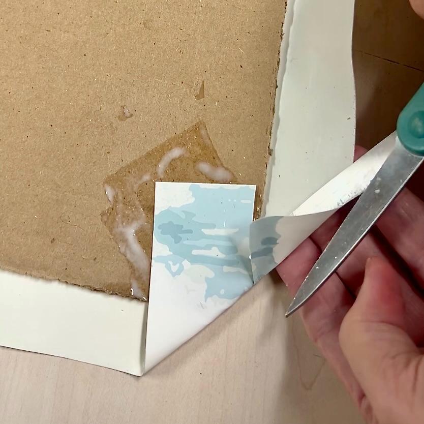 A hand uses scissors to lift and peel the edge of a print from a cardboard backing, much like removing inserts from a Functional Basket Tray. The corner is partially separated, revealing adhesive and residue underneath.
