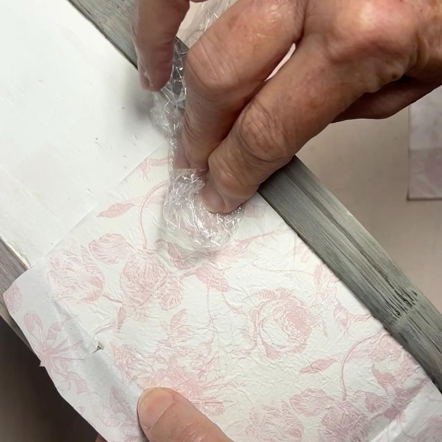A person presses crumpled plastic wrap onto a surface covered with pink floral-patterned paper and masking tape, next to a strip of gray-painted wood—perfect inspiration for your next thrifted basket makeover.