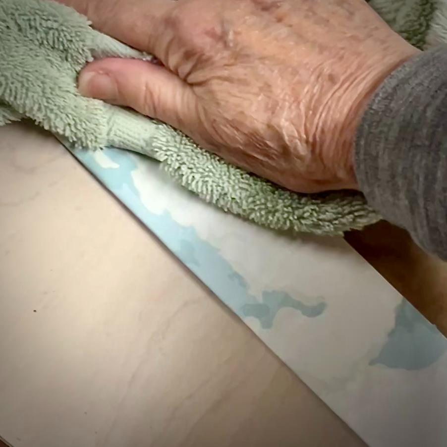 A hand holding a green cloth wipes a light wooden surface near a Functional Basket Tray and a strip of wallpaper featuring a blue and white pattern.