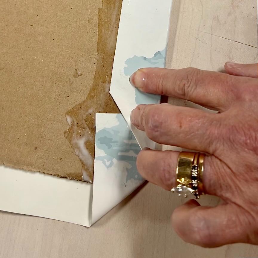 A hand with gold rings folds and presses the corner of a blue-patterned paper onto the back of a Functional Basket Tray, using glue to secure it. Some glue is visible on both surfaces as the paper adheres smoothly.