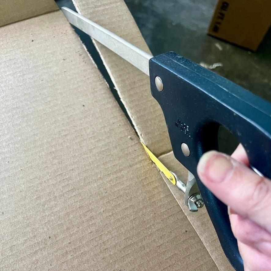 A hand uses a hacksaw with a black handle to cut through a large piece of cardboard, shaping it into a functional basket tray.