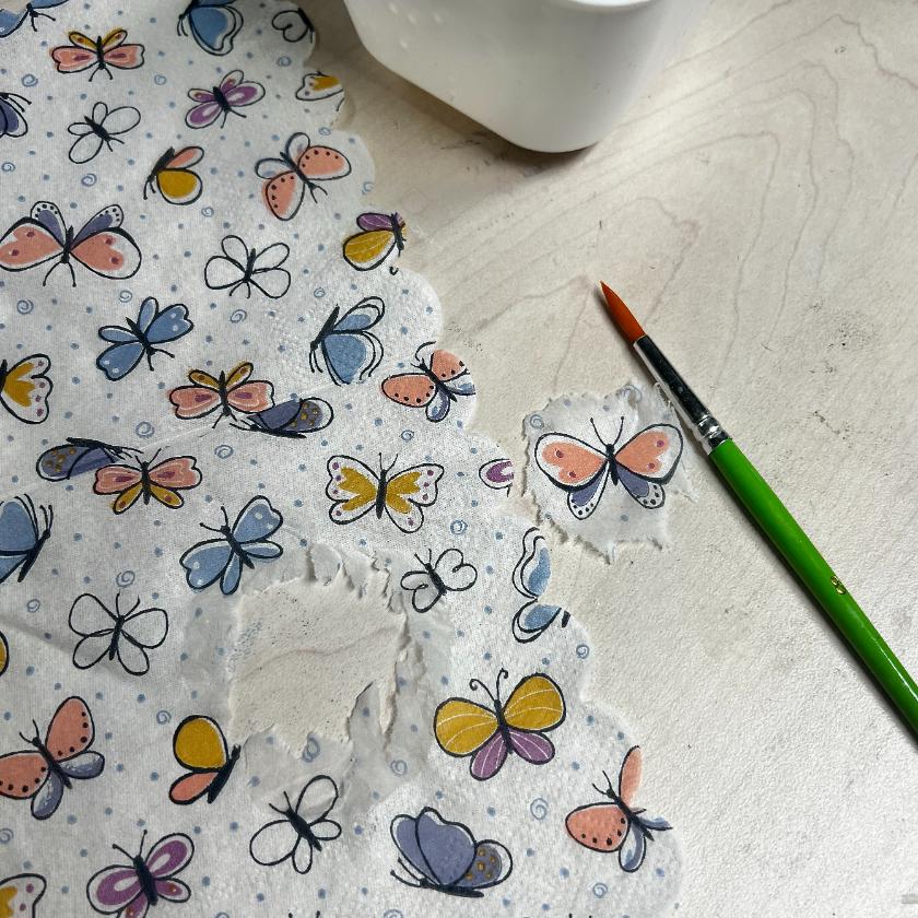 A sheet of tissue paper with colorful butterfly prints is laid on a light wooden surface, perfect for a thrifted basket makeover. Part of the paper is torn, and a paintbrush with a green handle lies nearby next to a white container.