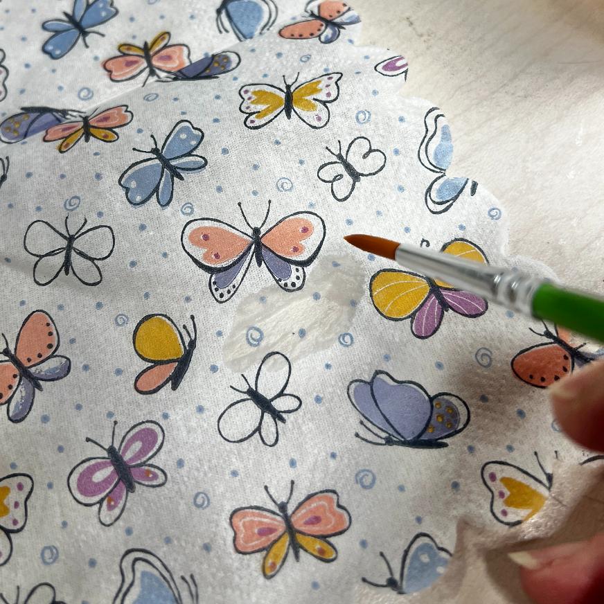 A hand uses a paintbrush to apply clear liquid onto white fabric with colorful butterfly patterns and scalloped edges during a thrifted basket makeover.