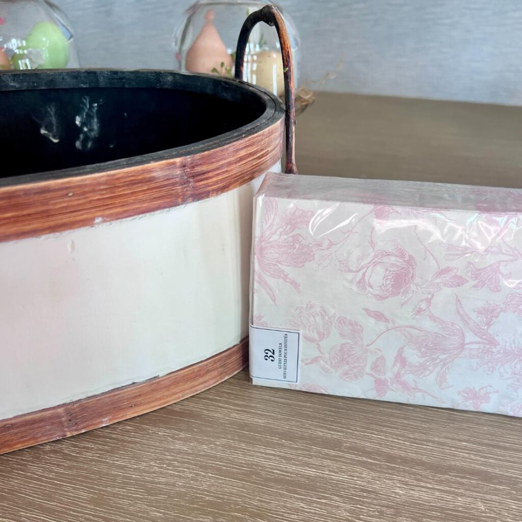 A rectangular wooden basket, refreshed with a thrifted basket makeover and a black interior, sits next to pink floral-patterned napkins on a wooden table. In the background, pastel-colored decorative eggs are visible.