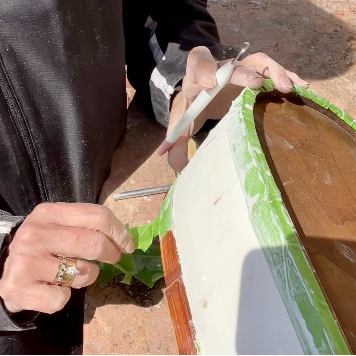A person peels green painter’s tape from the edge of a wooden surface during a thrifted basket makeover, holding a sculpting tool as sunlight casts shadows on their hands. The surface beneath is partially painted white.