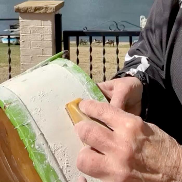 A person sands the side of a thrifted basket makeover, covered in white paint or primer and edged with painter's tape. A lake and yard are visible in the background.