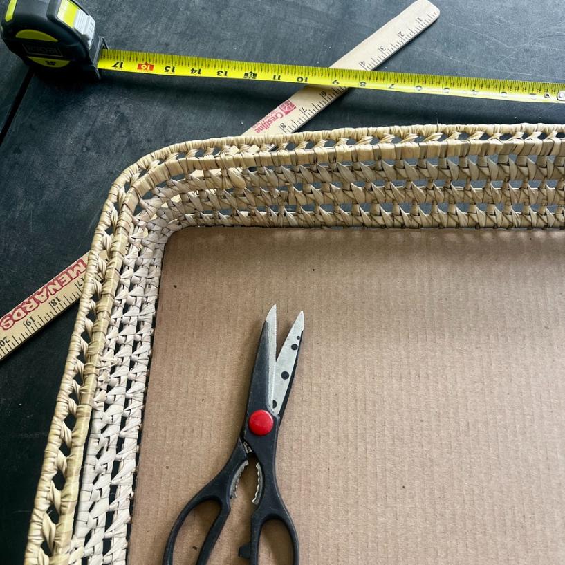 A functional basket tray sits on a table with a pair of scissors inside it. A tape measure and a wooden ruler are placed nearby, partially overlapping at the top of the image.