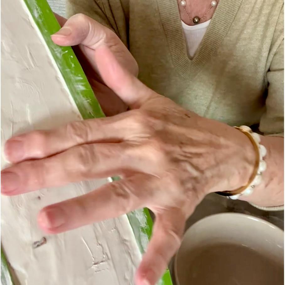A person wearing a beige sweater and jewelry smooths white plaster onto a green-edged surface with their hand during a thrifted basket makeover; a bowl sits on the table nearby.