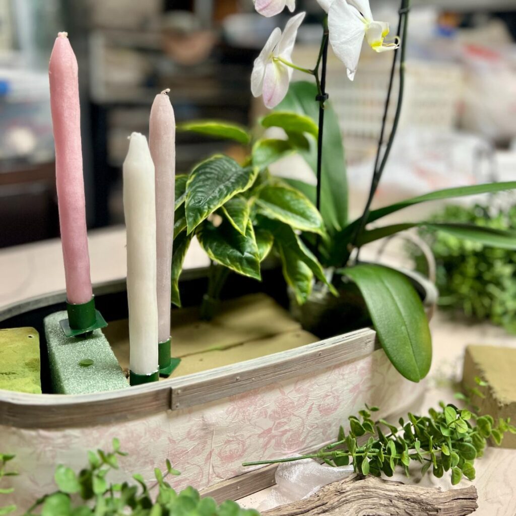 A close-up of a floral arrangement in a thrifted basket makeover, featuring two pastel taper candles, artificial plants, and an orchid, surrounded by green foliage and floral foam blocks on a table.