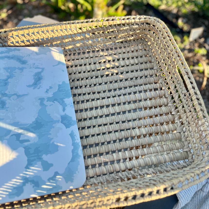 A close-up of a functional basket tray woven from wicker, with a blue and white patterned item, possibly a notebook, partially inside it. The tray is placed outdoors, where sunlight casts intricate shadows.