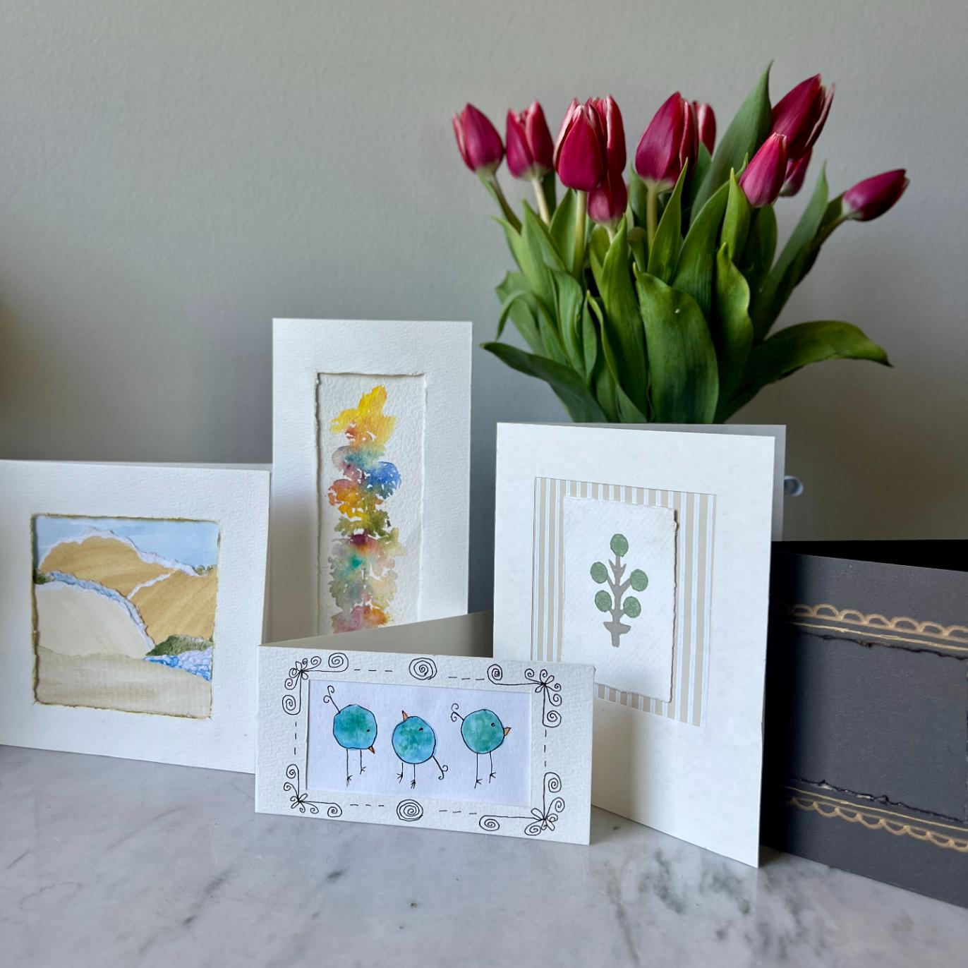 Five handmade greeting cards with various designs are displayed on a marble surface in front of a vase of pink tulips. The cards feature abstract art, floral patterns, and whimsical blue birds.