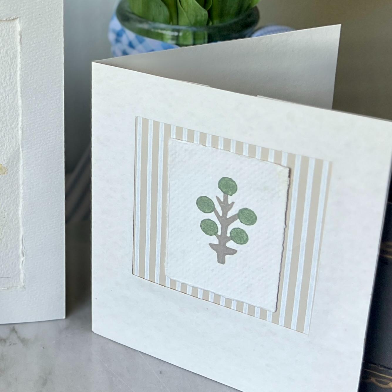 A white greeting card with a simple tree illustration featuring a brown trunk and green round leaves, framed by vertical beige and white stripes, standing upright on a surface.