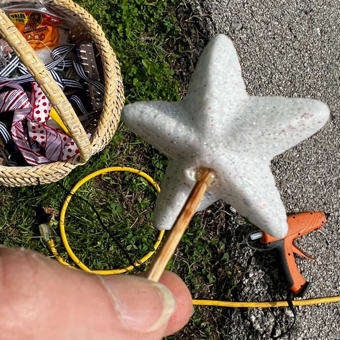 A close-up of a hand holding a white star-shaped object on a stick, perfect for crafting a DIY patriotic mailbox swag. In the background: ribbon spools in a basket, an orange hot glue gun, yellow extension cord, grass, and pavement.