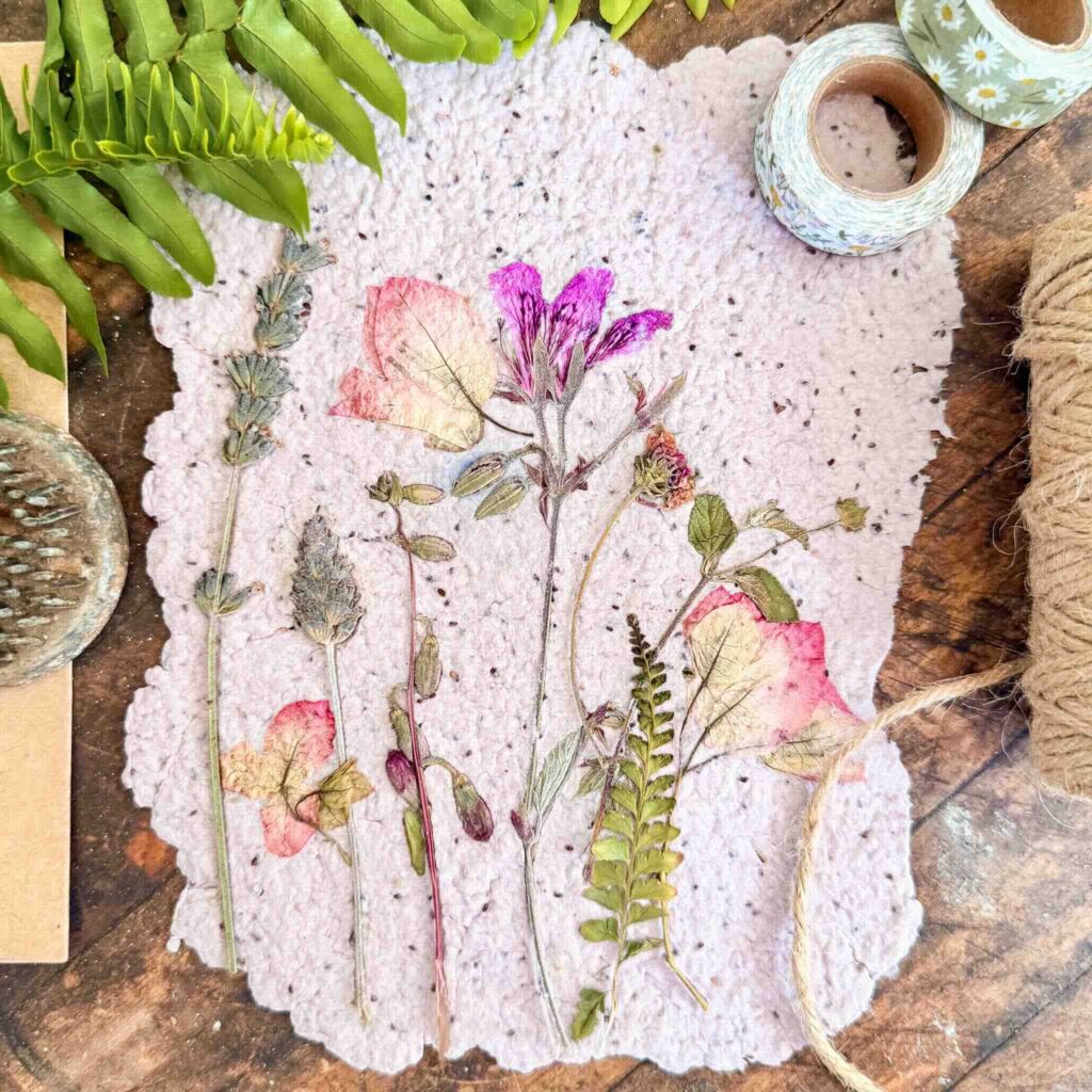 A handmade sheet of paper with pressed flowers and ferns arranged on top, surrounded by washi tape, twine, a fern leaf, and a brush on a wooden surface.