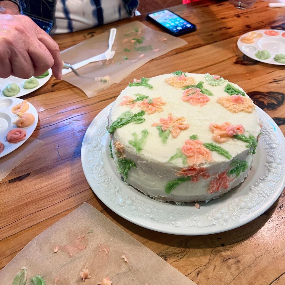 A round white cake decorated with pink and orange flower designs sits on a plate on a wooden table. Nearby are paint palettes with icing, a phone, and a hand holding a fork next to parchment paper.