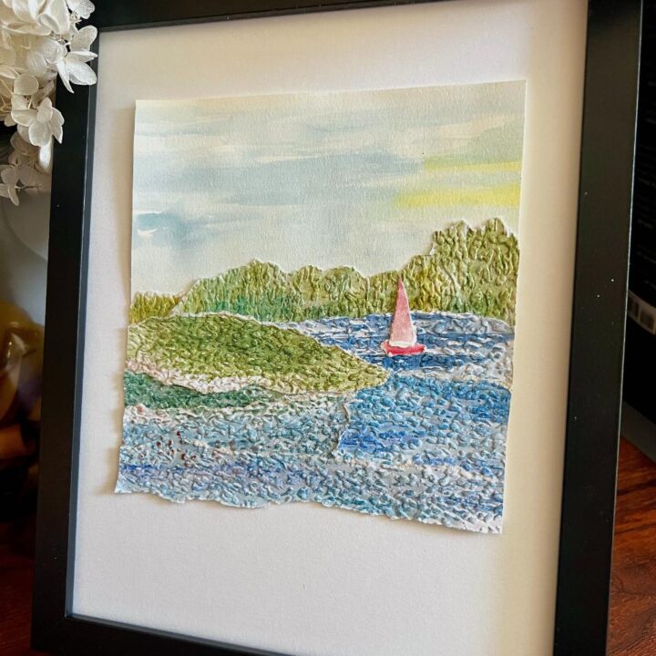 Framed textured artwork of a small sailboat with a red sail on blue water, surrounded by green hills and trees, under a lightly clouded sky. The art is displayed in a black frame on a wooden surface.