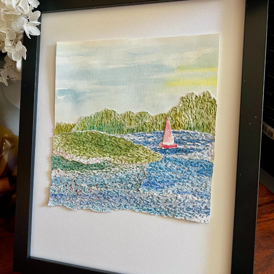 Framed textured artwork of a small sailboat with a red sail on blue water, surrounded by green hills and trees, under a lightly clouded sky. The art is displayed in a black frame on a wooden surface.