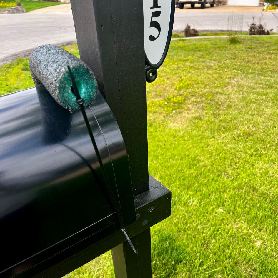 A pool noodle piece, possibly part of a DIY patriotic mailbox swag, is attached to the edge of a black mailbox with a zip tie as a protective buffer. Green grass and a street are visible in the background.
